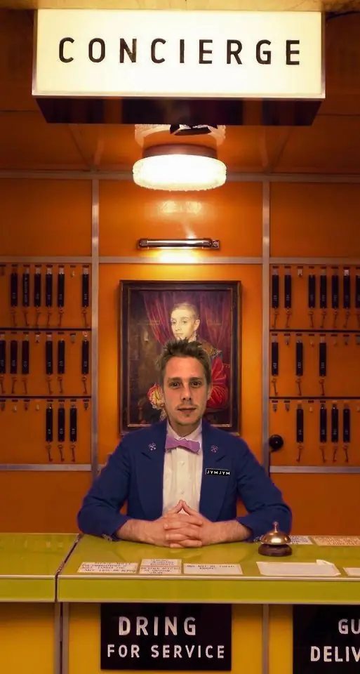Portrait du fondateur de JymJym Conciergerie assis en costume avec nœud papillon dans un décor inspiré du film Grand Budapest Hotel, illustrant l'élégance et le professionnalisme du service de conciergerie en Guadeloupe.